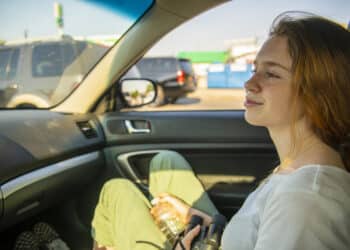 The attractive long-haired Caucasian young girl resting at the passenger seat in the car during the roadtrip in the USA. Summer vacations, the sunny hot day on the city street.