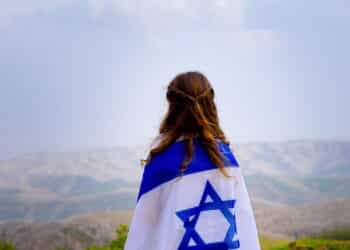 Little patriot jewish girl standing and enjoying great view on the sky, valley and mountains with the flag of Israel wrapped around her. Memorial day-Yom Hazikaron and Yom Ha'atzmaut concept.