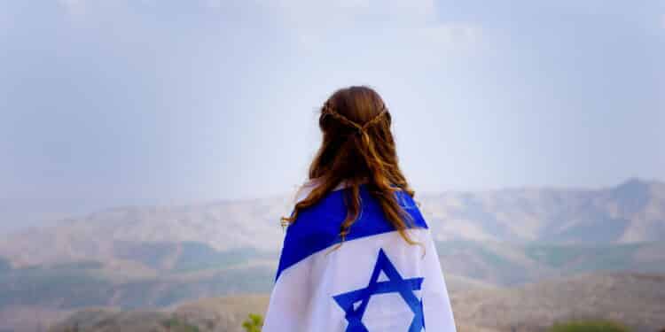 Little patriot jewish girl standing and enjoying great view on the sky, valley and mountains with the flag of Israel wrapped around her. Memorial day-Yom Hazikaron and Yom Ha'atzmaut concept.