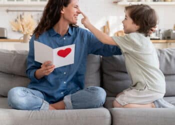 Joyful mom in playful mood laughing cuddle with cute little son hold gift on birthday sitting on couch at home. Preschool boy congratulating mum on mothers day, presenting greeting card with red heart