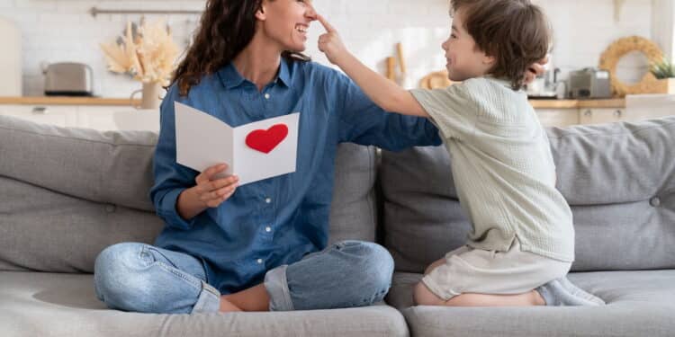 Joyful mom in playful mood laughing cuddle with cute little son hold gift on birthday sitting on couch at home. Preschool boy congratulating mum on mothers day, presenting greeting card with red heart