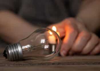 An incandescent light bulb and a candle in the hands of an old woman on a wooden table, people without light, the light of a candle when there is no light in Ukraine