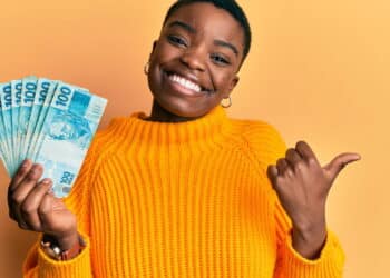 Young african american woman holding 100 brazilian real banknotes pointing thumb up to the side smiling happy with open mouth