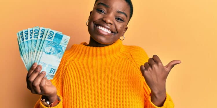 Young african american woman holding 100 brazilian real banknotes pointing thumb up to the side smiling happy with open mouth