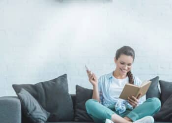 Mulher lendo livro enquanto liga o ar condicionado com controle remoto em casa - Créditos: depositphotos.com / AllaSerebrina