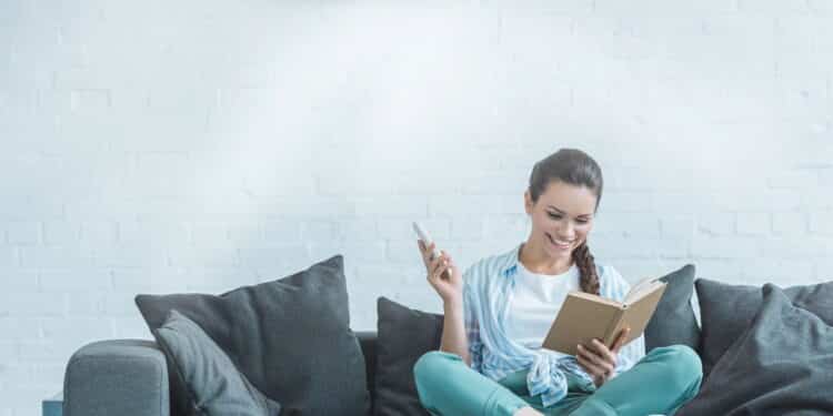 Mulher lendo livro enquanto liga o ar condicionado com controle remoto em casa - Créditos: depositphotos.com / AllaSerebrina