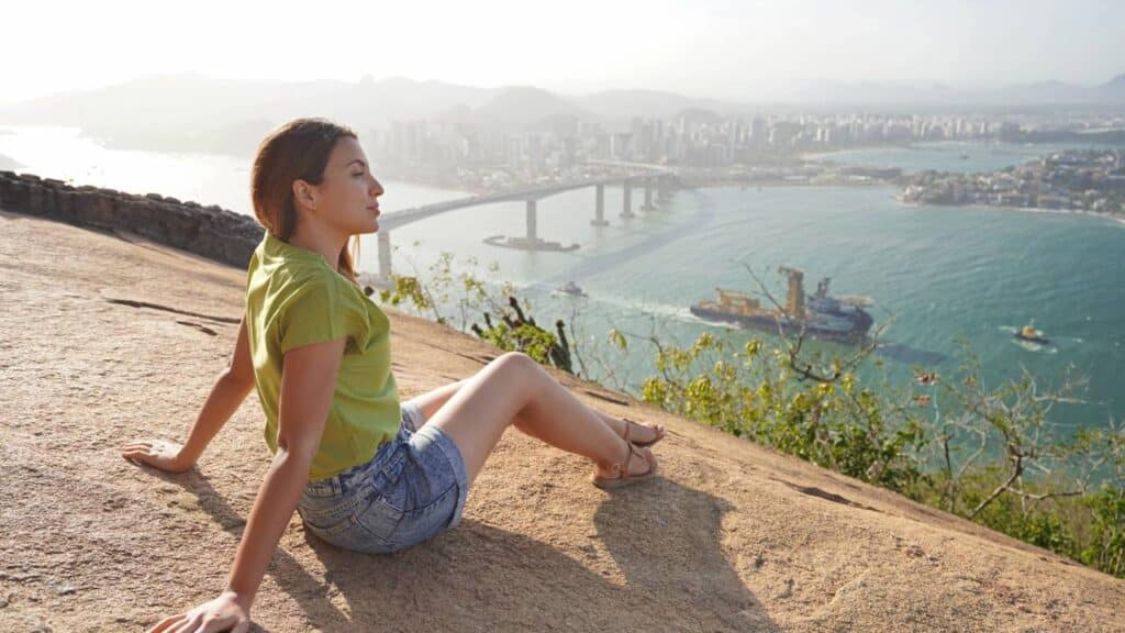 Linda mulher sentada relaxadamente apreciando a paisagem no topo da montanha ao pôr do sol - Créditos: depositphotos.com / sergio_pulp