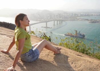Linda mulher sentada relaxadamente apreciando a paisagem no topo da montanha ao pôr do sol - Créditos: depositphotos.com / sergio_pulp