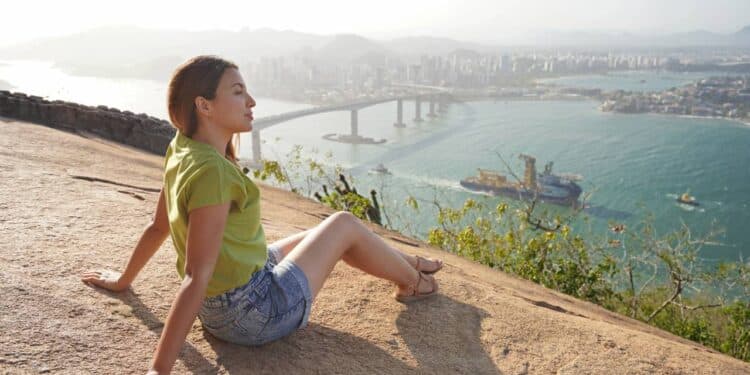 Linda mulher sentada relaxadamente apreciando a paisagem no topo da montanha ao pôr do sol - Créditos: depositphotos.com / sergio_pulp