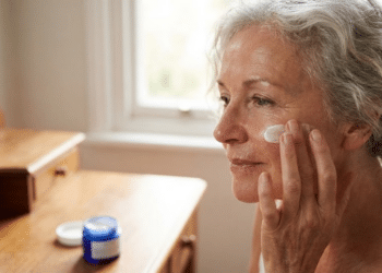 radiante de mulher 60+ com pele hidratada aplicando creme no rosto, com potes de marcas populares desfocados na frente