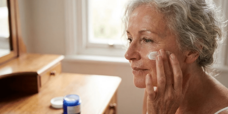 radiante de mulher 60+ com pele hidratada aplicando creme no rosto, com potes de marcas populares desfocados na frente