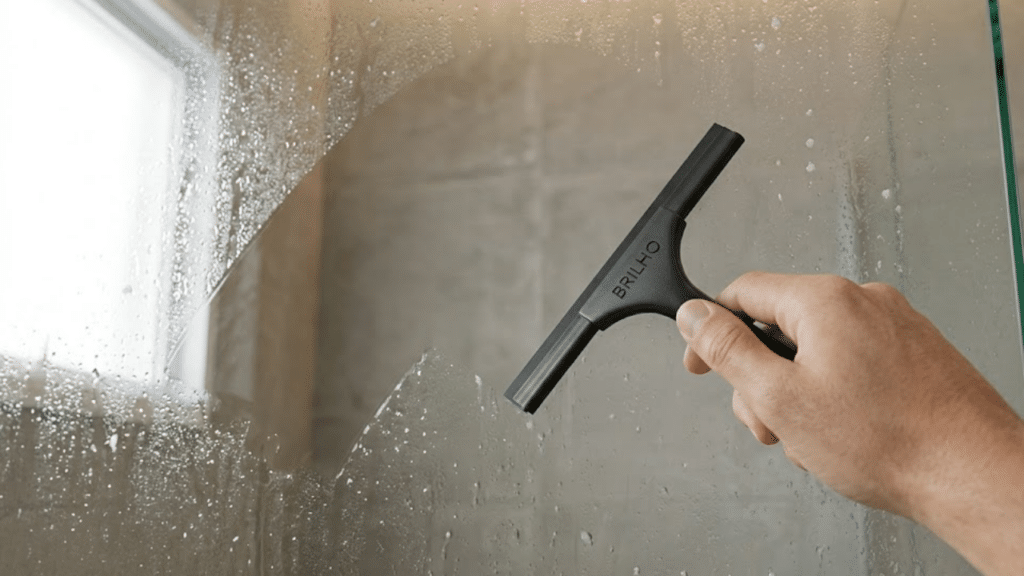 Mão passando rodo no vidro molhado, revelando superfície límpida