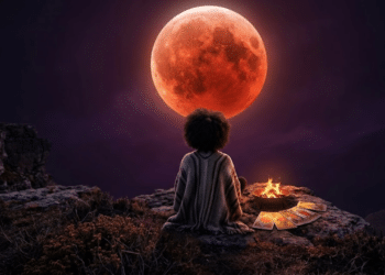 Uma pessoa de costas em um penhasco rochoso, observando a imponente Lua de Sangue no céu noturno ao lado de uma fogueira e cartas de tarô.