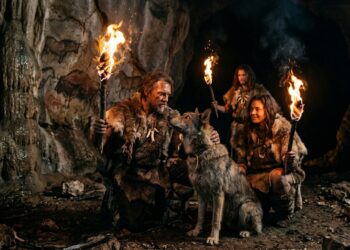 Grupo de humanos pré-históricos segura tochas dentro de caverna, ao lado de um cão, com pinturas rupestres nas paredes.