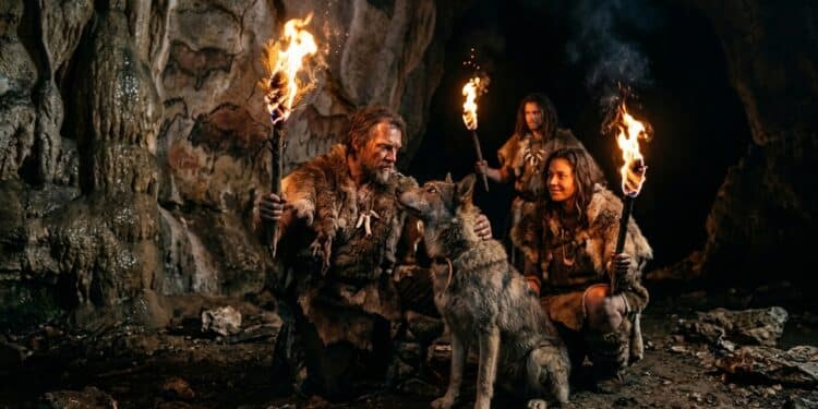 Grupo de humanos pré-históricos segura tochas dentro de caverna, ao lado de um cão, com pinturas rupestres nas paredes.