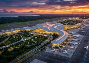 Vista aérea de um aeroporto moderno ao entardecer, com terminal iluminado em formato curvo, aviões estacionados, pistas extensas e floresta ao redor.