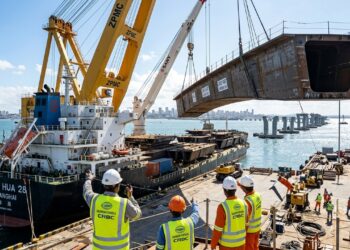 Guindastes gigantes descarregam uma grande estrutura metálica de um navio cargueiro, com trabalhadores de capacete acompanhando a operação no porto.