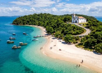 Vista aérea de praia paradisíaca com águas cristalinas, barcos ancorados, faixa de areia clara, vegetação preservada e igreja histórica no alto.