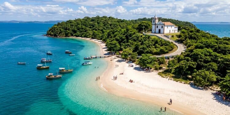 Vista aérea de praia paradisíaca com águas cristalinas, barcos ancorados, faixa de areia clara, vegetação preservada e igreja histórica no alto.