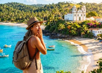 Turista observa praia de águas cristalinas no Nordeste, com barcos, vegetação tropical e igreja histórica ao fundo.